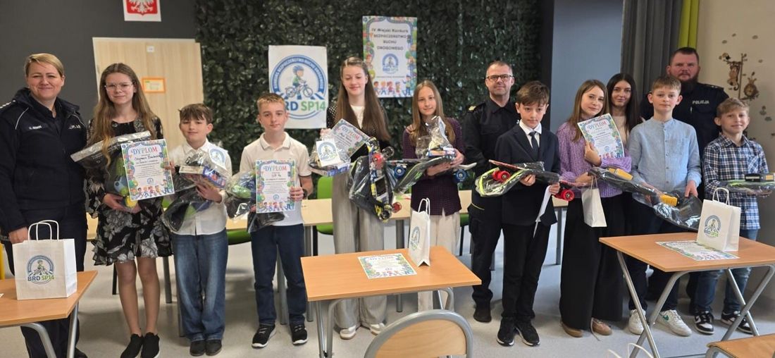 Laureaci IV Miejskiego Konkursu BRD pozują z dyplomami i nagrodami (kaskami, akcesoriami). Obok nich stoją uśmiechnięci policjanci i strażnik miejski w sali SP nr 14 w Jastrzębiu-Zdroju