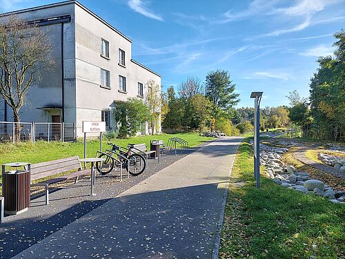 Stacja obsługi i odpoczynku dla rowerzystów przy ścieżce rowerowej. Wzdłuż asfaltowej ścieżki ustawione są drewniane ławki, kosze na śmieci, stojaki z zaparkowanymi rowerami oraz tablica informacyjna. Po lewej stronie widać jasny, dwupiętrowy budynek