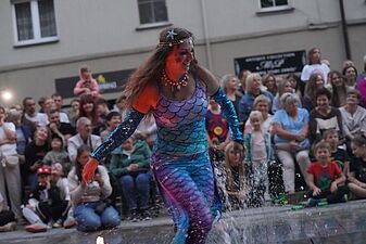 Aktorka w kolorowym stroju z motywem łusek (syrena) tańczy dynamicznie, rozbryzgując wodę w podświetlonej fontannie, przed publicznością na placu miejskim
