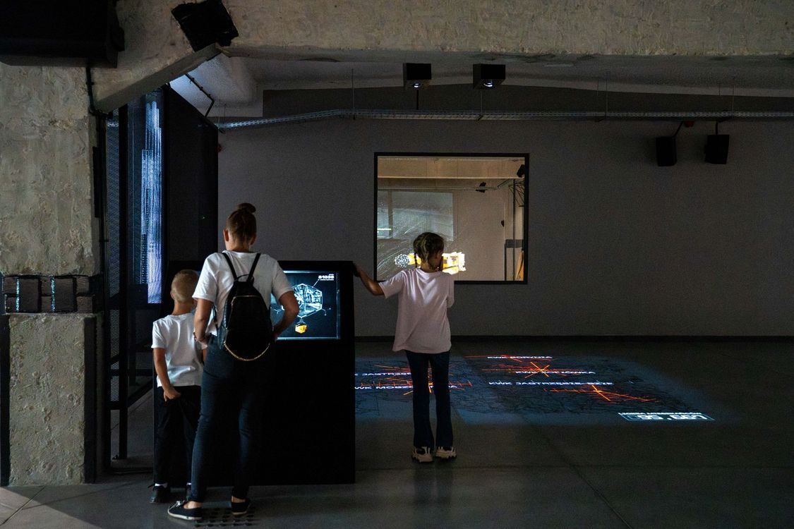 Dzieci korzystające z multimedialnych ekranów i interaktywnej mapy wyświetlanej na podłodze w ciemnej sali wystawowej