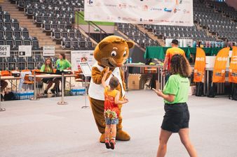 Człowiek w przebraniu maskotki wita się z maych chopcem, obok przechodząca kobieta, w tle widok na krzesełka hali sportowej