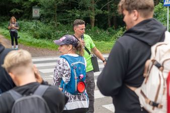 Organizator imprezy w jaskrawozielonej koszulce mówi do mikrofonu, stojąc na przejściu dla pieszych obok uczestników z psami
