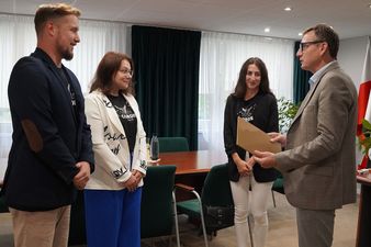 Prezydent Michał Urgoł wręcza brązową teczkę z dokumentami trzem osobom stojącym przed nim w biurze. Twoje rozmówczynie mają czarne koszulki z napisem „teatr CORDIS”