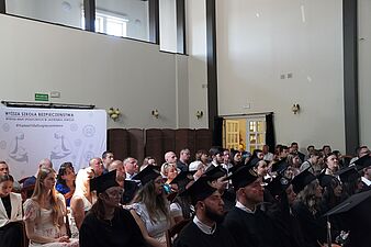 Uczestnicy i absolwenci w biretach siedzący w ławkach podczas inauguracji lub graduacji Wyższej Szkoły Bezpieczeństwa