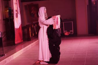 Klęczący aktor utożsamiający postać Księdza Machy, za Nim stojąca kobieta ubrana na biało trzyma przed Nim w rękach chustę z wizerunkiem Chrystusa