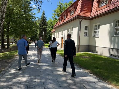 Grupa osób idzie kamienną alejką wzdłuż bocznej elewacji budynku. Wokół rosną wysokie drzewa