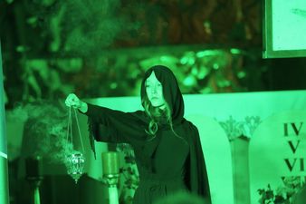 Kobieta ubrana na czarno trzyma w rękach kadzidło, wszystko podświetlone zielonym światłem