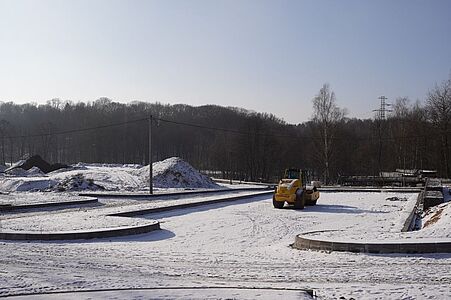 Szeroki, ośnieżony plac budowy drogi, po którym porusza się żółty walec drogowy
