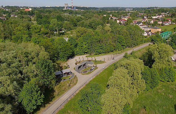 Szerokie ujęcie z drona przedstawiające trasę rowerową i miejsce obsługi rowerzystów na tle panoramy miasta, otaczających zielonych terenów, domów jednorodzinnych oraz zabudowań przemysłowych w oddali