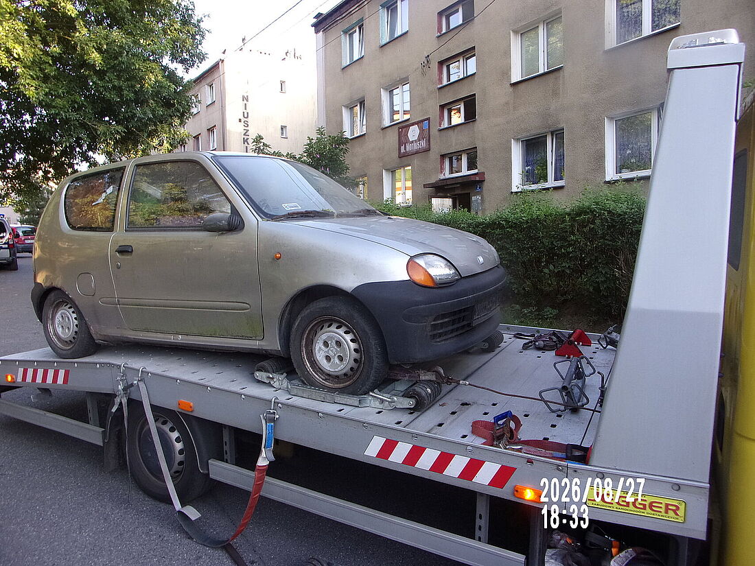 Srebrny Fiat Seicento przymocowany pasami do platformy żółtej lawety holowniczej. W tle widać wielopiętrowy budynek mieszkalny z tabliczką adresową ulicy Moniuszki