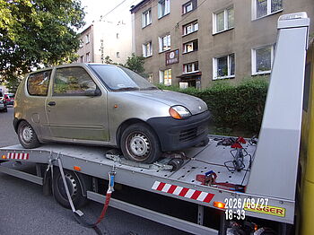 Srebrny Fiat Seicento przymocowany pasami do platformy żółtej lawety holowniczej. W tle widać wielopiętrowy budynek mieszkalny z tabliczką adresową ulicy Moniuszki