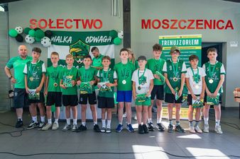 Kolejna drużyna zawodników w zielonych koszulkach wraz z trenerem, prezentująca zdobyte medale i otrzymane upominki