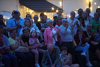 Licznie zgromadzona publiczność w różnym wieku ogląda i oklaskuje plenerowy występ artystyczny o zmierzchu