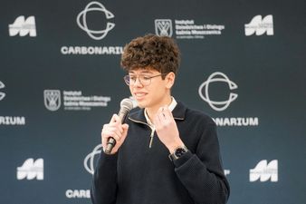 chłopak w ciemnych włosach z okularami trzyma w dłoni mikrofon i debatuje