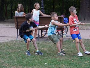 Trzech chłopców z zaangażowaniem przeciąga linę na trawie w parku, a w tle na ławce siedzi dziewczynka i dorosła osoba