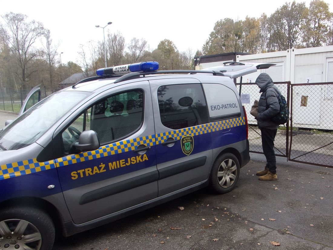 Zdjęcie przedstawia ludzi przy samochodzie straży miejskiej