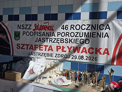 Baner informacyjny dotyczący sztafety pływackiej z okazji 46. rocznicy podpisania Porozumienia Jastrzębskiego, powieszony nad stołem z pucharami i nagrodami