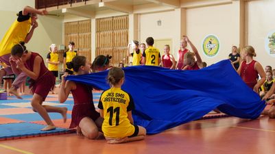 Uczniowie prezentujący występ artystyczny z użyciem dużej płachty niebieskiego materiału na hali sportowej