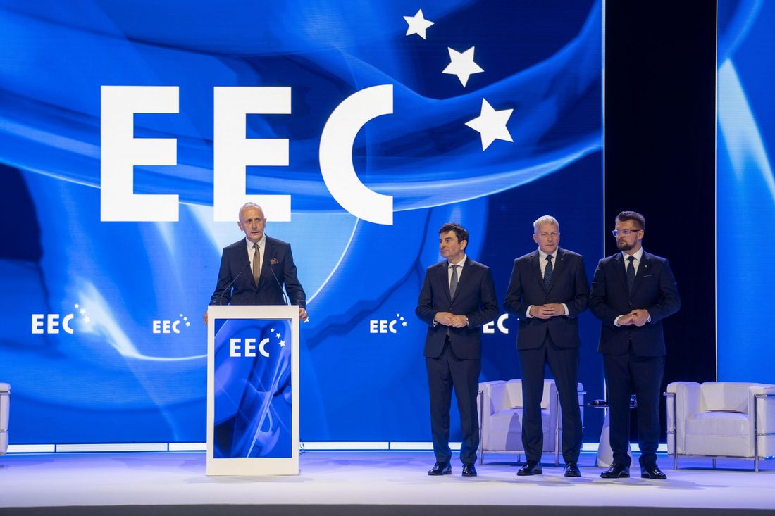 Czterech mężczyzn w garniturach na scenie w tle ekran z napisem EEC i trzema białymi gwiazdami