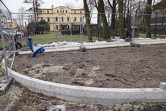 Widok na wyprofilowany, półokrągły fragment terenu pokryty ziemią i oddzielony nowymi, granitowymi krawężnikami, zabezpieczony metalowym płotem. W tle za płotem znajduje się trawnik i zabytkowy, żółty budynek hotelu Dąbrówka