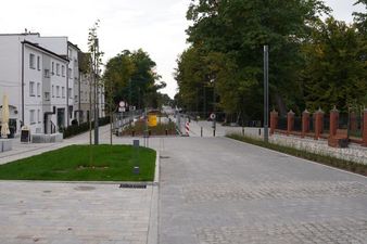 Szeroki kadr przedstawiający perspektywę nowej promenady miejskiej. Po lewej stronie widoczne są jasne elewacje budynków, po prawej park z ceglanym ogrodzeniem, a na pierwszym planie niewielki trawnik