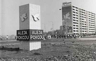 Tablica wjazdowa/pamiątkowa "Aleja Pokoju" z gołębiami, w tle duży blok z muralem i napis "osiedle Morcinka"