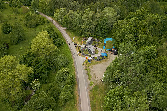 Widok z drona na ścieżkę rowerową i przyległą strefę rekreacyjną z niebieskim torem pumptrack, wiatami oraz namiotami eventowymi, otoczoną gęstym lasem