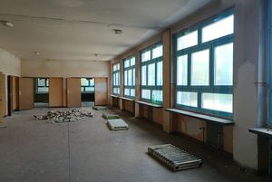 Szeroki, pusty korytarz w trakcie remontu. Wzdłuż ściany z oknami ciągną się długie blaty. Na podłodze leżą zdemontowane kaloryfery oraz sterta gruzu i papierów