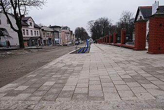 Ujęcie przedstawia nieutwardzoną, gruntową drogę biegnącą wzdłuż białego budynku kościoła lub klasztoru z dużą rzeźbą ukrzyżowanego Chrystusa na fasadzie. Przed budynkiem budowany jest nowy chodnik, oddzielony od jezdni pasem ziemi
