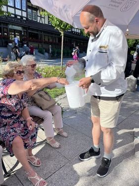 Mężczyzna w stroju kucharskim częstuje dwie siedzące kobiety produktem z białego pojemnika podczas plenerowego wydarzenia