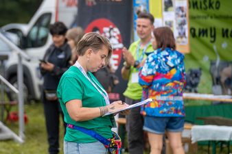 Kobieta w zielonej koszulce organizatora zapisuje dane na podkładce z klipsem; w tle rozmawiają inne osoby na festynie