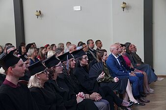 Absolwenci w czarnych togach i biretach oraz zaproszeni goście siedzą w rzędach na widowni podczas uroczystości akademickiej