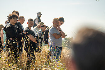 Grupa widzów stojąca w wysokiej trawie i obserwująca przejazd aut rajdowych