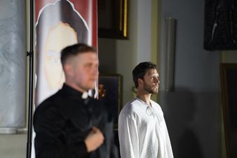 Mężczyzna ubrany na biały uosobiający Jezusa Chrystusa, na pierwszym planie mężczyzna ubrany w sutannę utożsamiający księdza Jana Machy