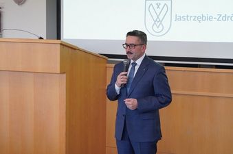 Roman Cichowicz w garniturze przemawia do mikrofonu przy mównicy. W tle ekran z logo i napisem „Jastrzębie-Zdrój
