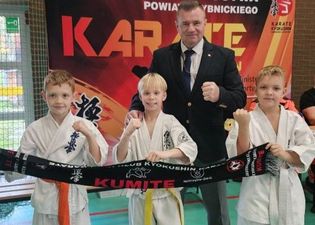 Najmłodsi zawodnicy Jastrzębski Klub Kyokushin Karate Kumite pozujący do zdjęcia z trenerem i trzymający rozciągniety szalik klubowy