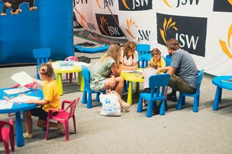 Dzieci i dorośli siedzą przy małym stoliczku, obok inna dziewczynka siedząca samotnie rysująca obrazek kredkami