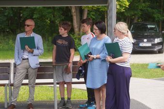 Grupa kilku osób, w tym mężczyzna w jasnej marynarce, dwóch nastolatków i dwie kobiety, stoi na zewnątrz i patrzy w jedną stronę. Mężczyzna trzyma folder, a jedna z kobiet dyplom