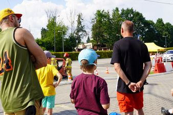 Widzowie, w tym dorośli i dzieci w letnich ubraniach, stoją tyłem i obserwują z bliska pokaz akcji ratowniczej straży pożarnej na placu