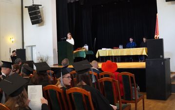 Absolwenci w togach i biretach siedzą na widowni. W tle na scenie widoczne są władze uczelni, w tym rektor dr Andrzej Zduniak przy stole prezydialnym