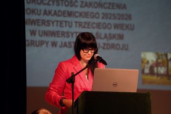 Kobieta profesor Kornelia Batko z czarnymi włosami w okularach i różowym stroju przy ambonie przemawia ze sceny w tle ekran
