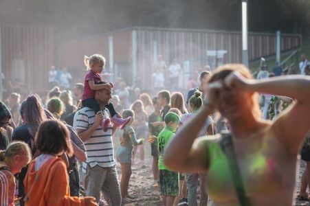 Rodziny z dziećmi bawiące się w chmurze pyłu na festiwalu kolorów; dziewczynka siedzi na barankach u taty