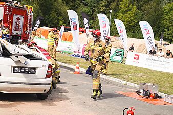 Strażak w pełnym umundurowaniu ochronnym i czerwonym hełmie idzie w stronę kamery obok rozbitego białego auta; na ziemi leżą narzędzia hydrauliczne, w tle widoczne są flagi reklamowe i namioty