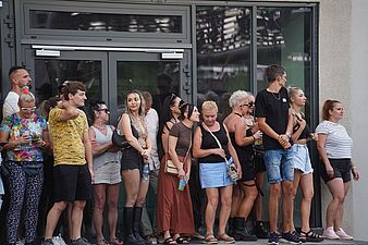 Grupa ludzi stojąca pod oszklonym wejściem do budynku i obserwująca imprezę plenerową