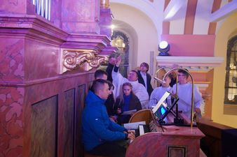 Osoba grająca na organach i śpiewający ludzie. W tle duchowny święci organy