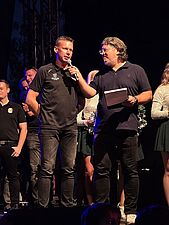 Rafał Bernacki przemawia do mikrofonu trzymanego przez prowadzącego na scenie podczas imprezy plenerowej