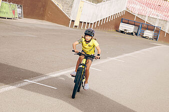 Dziewczynka w kasku i żółtej koszulce jedzie na rowerze po torze wokół boiska sportowego