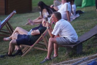 Mężczyzna siedzący na drewnianej ławie fotografuje smartfonem wydarzenie festiwalowe. W tle na zboczu inni ludzie wypoczywają na leżakach