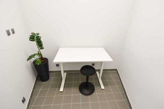 Małe pomieszczenie biurowe z białym biurkiem i czarnym hokerem. Obok stoi wysoka roślina doniczkowa (zamiokulkas). Jasne ściany i szara podłoga z płytek. Minimalistyczny wystrój