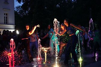 Aktorzy w mieniących się strojach wykonują układ choreograficzny pośród tryskających strumieni podświetlonej fontanny